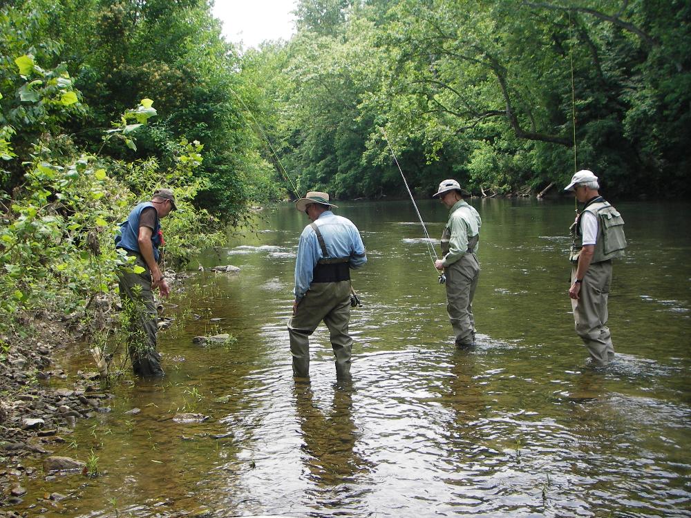 Three student anglers learning about minnows and where they are found in the river from Dale, the instructor all four are standing in a clear river carrying fly rods and wearing fishing waders and vests