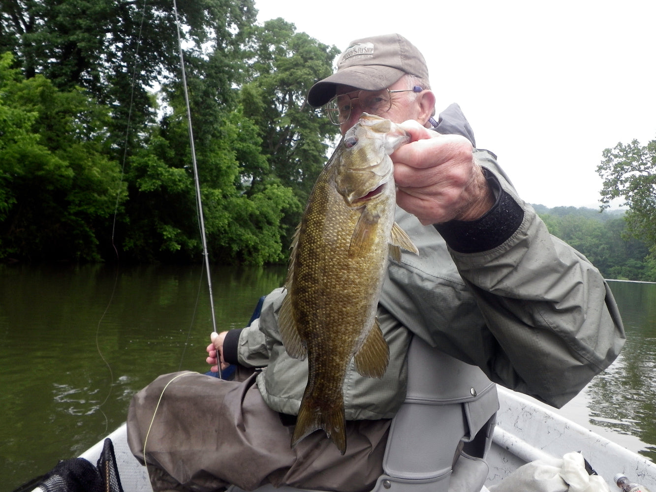 Harry Murray holding a nice smallmouth bass he caught on a fly rod in a Hyde drift boat on the Shenandoah River in Virginia. He is wearing a raincoat and fishing waders, holding a fly rod surrounded by water and green trees along the river bank