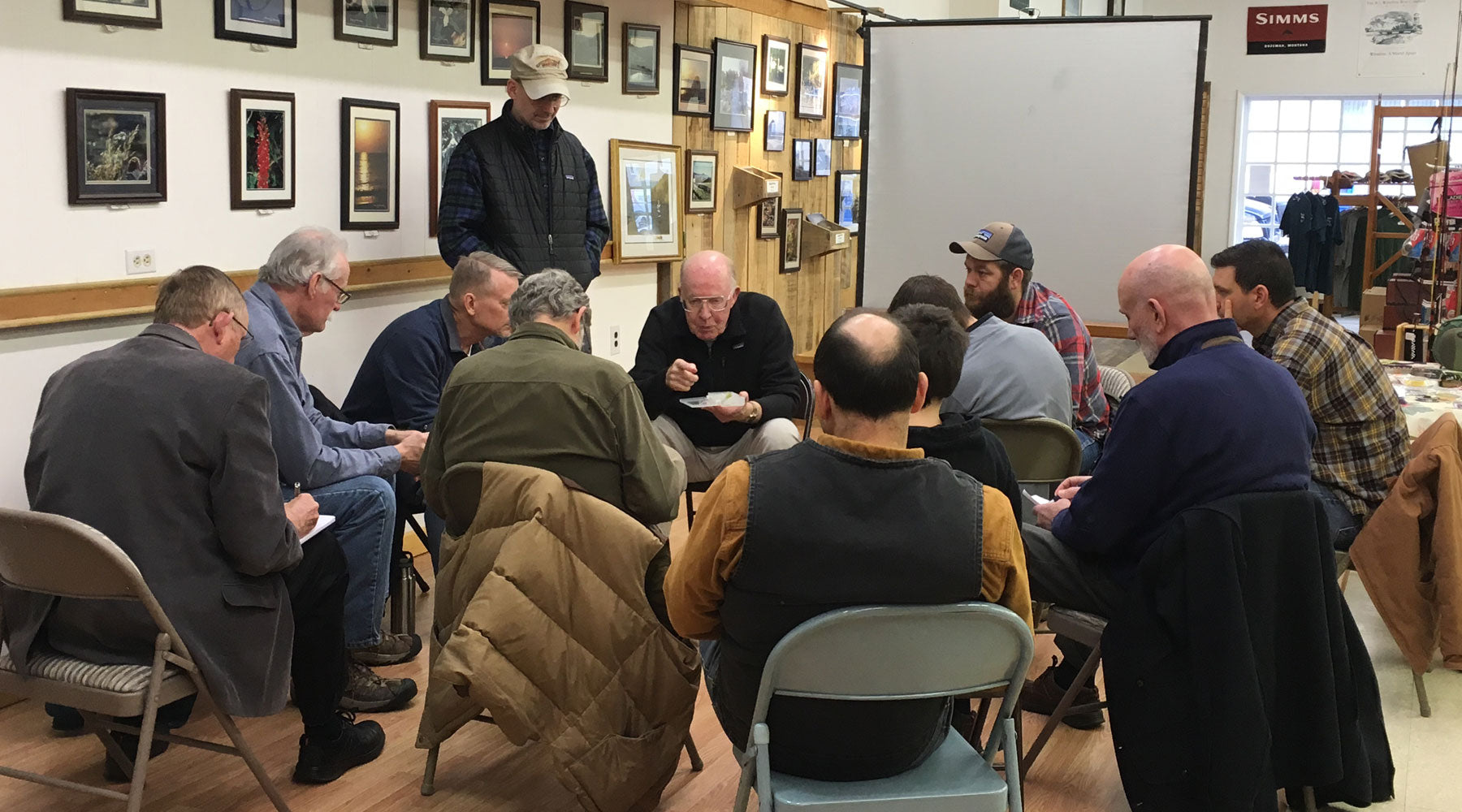 Harry Murray sitting on a chair explaining nymph selection while he is surrounded by students inside Murray's Fly Shop