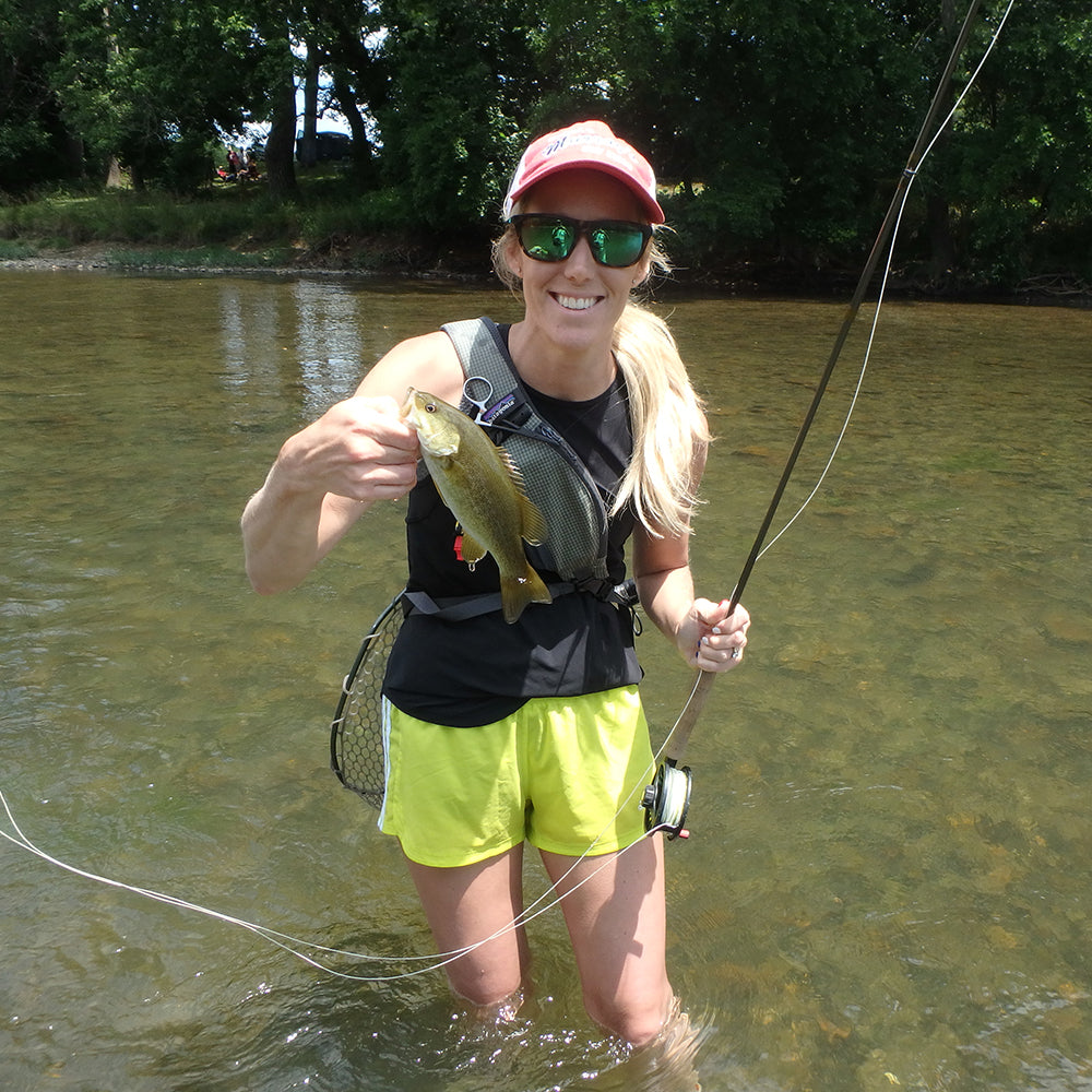 Shown is a girl standing in a river with green trees in the background. She is holding a fly rod and a smallmouth bass she caught on the fly