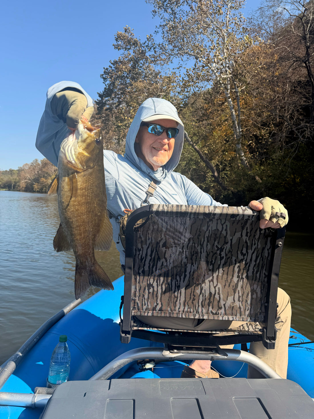 An anglers sitting in the front of a blue raft holding a large smallmouth bass with fall leaves on the trees in the background.