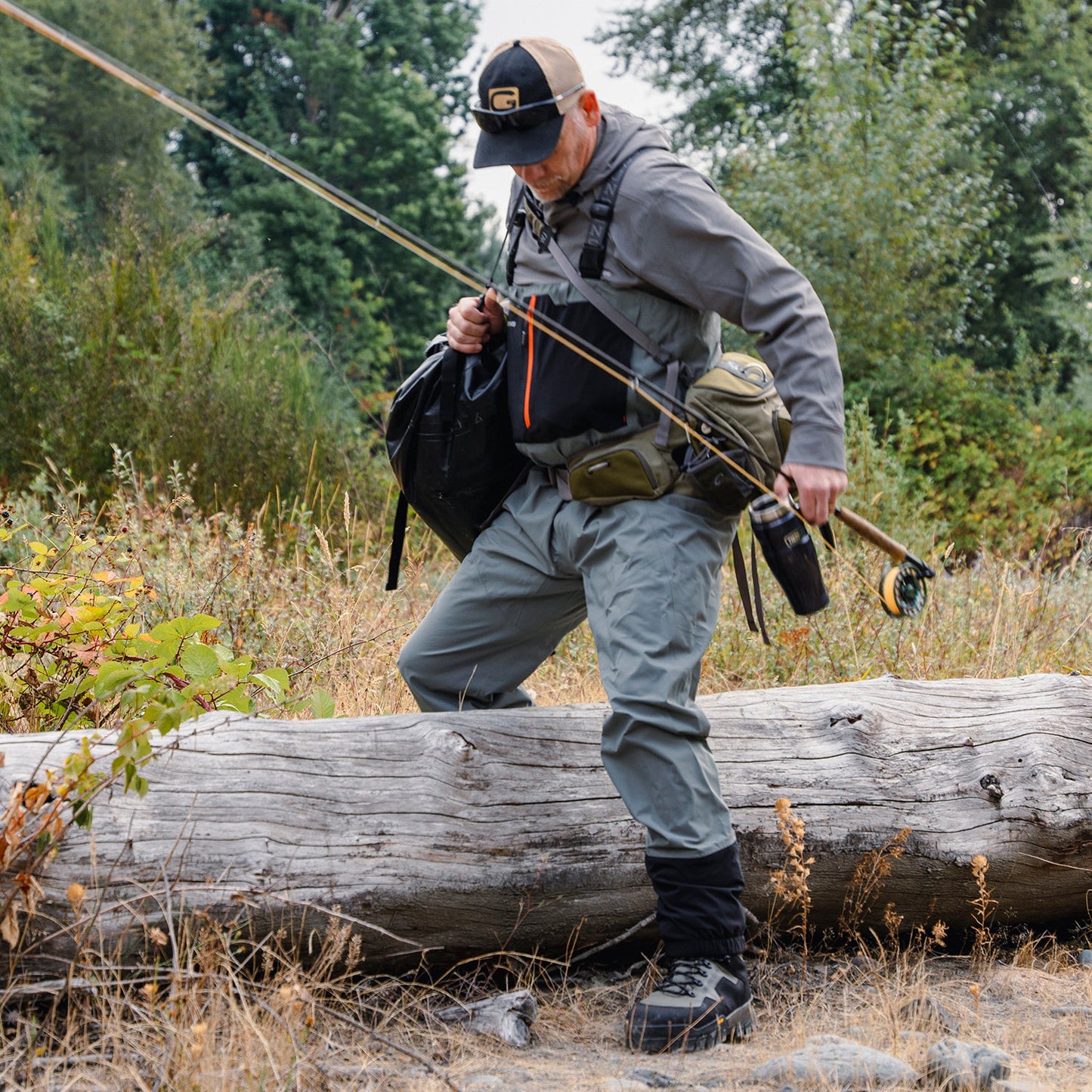 Grundens bedrock waders shown on an angler stepping over a log carrying a fly rod and wearing a fishing pack