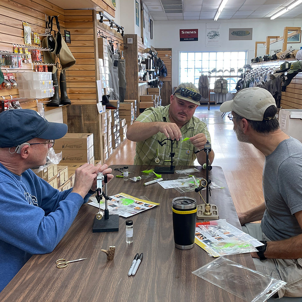 Minnow Changer Fly Tying Class with two students watching Brandon tie a segment in to the fly he is demonstrating on.  On the table are fly tying vises, instructions a various materials.  Picture is taken inside Murray's Fly Shop