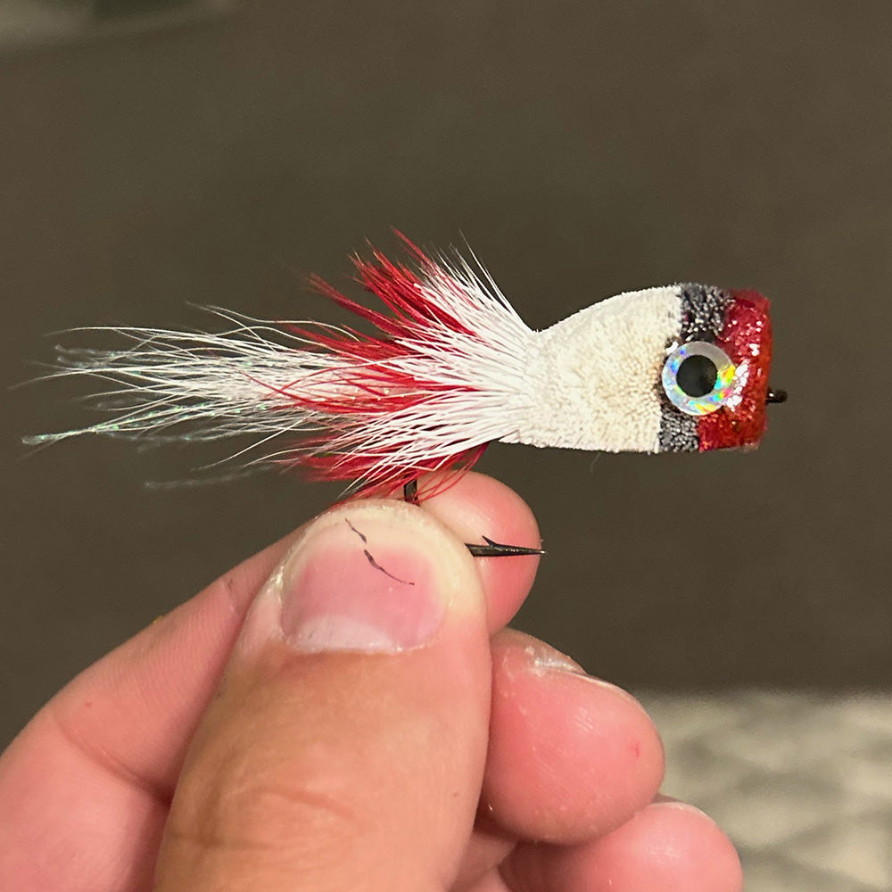 Up your deer hair bug tying skills with our fly tying class pictured is a red, gray and white deer hair bug being held by the tier