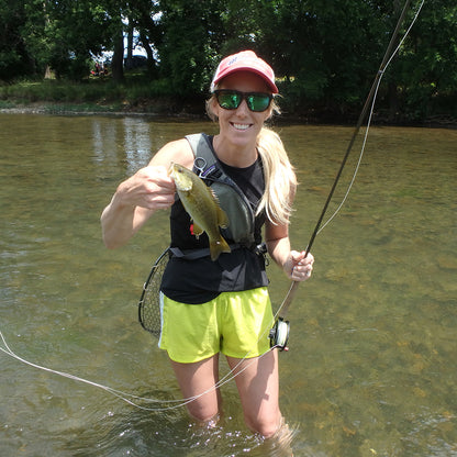 Shown is a girl standing in a river with green trees in the background.  She is holding a fly rod and a smallmouth bass she caught on the fly