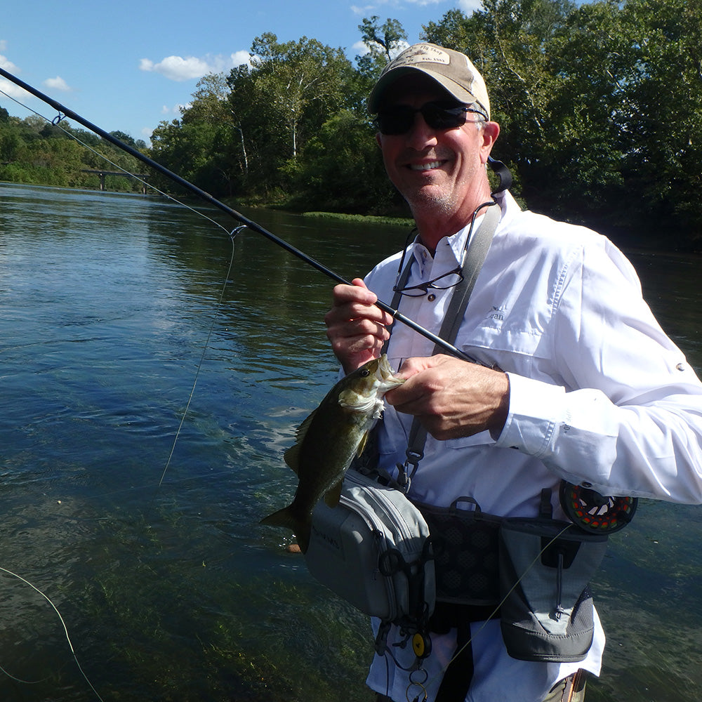 Shown is an angler in a white shirt holding a Smallmouth Bass and a fly rod standing in a river surrounded by green trees in a Murray's Fly Shop On The Stream Fly Fishing Schools