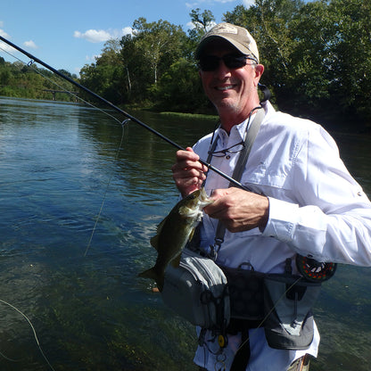 Shown is an angler in a white shirt holding a Smallmouth Bass and a fly rod standing in a river surrounded by green trees in a Murray's Fly Shop On The Stream Fly Fishing Schools